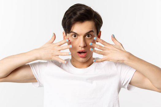 People, lgbtq and beauty concept. Close-up of beautiful gay man with glitter on face, showing nail polish on his fingernails, looking sassy at camera, standing over white background