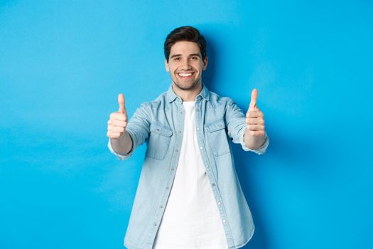 Smiling confident man showing thumbs up, guarantee quality, approving something good, standing against blue background