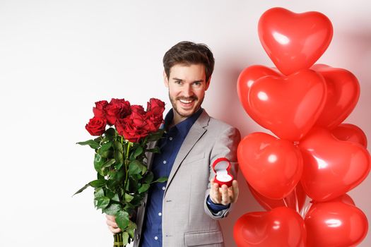 Handsome boyfriend in suit making a wedding proposal, showing engagement ring and say marry me, holding red roses, standing near Valentines day balloons, white background