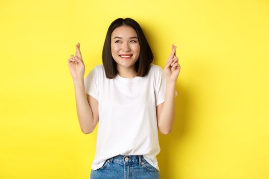 Hopeful asian girl smiling optimistic, feeling lucky, cross fingers and making wish, standing over yellow background