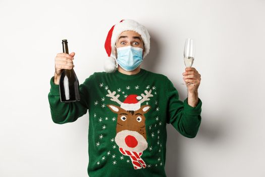 Concept of covid-19 and Christmas holidays. Happy man in face mask and Santa hat celebrating New Year with champagne, standing over white background