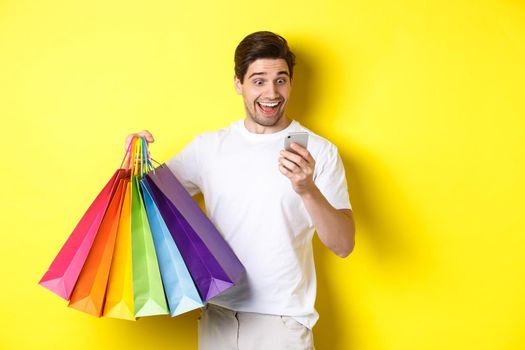 Excited man holding shopping bags and looking happy at mobile phone screen, standing over yellow background