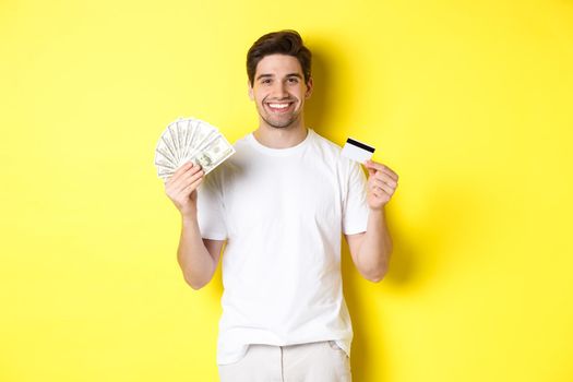 Young man withdraw money from credit card, smiling pleased, standing over yellow background