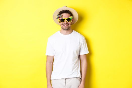 Concept of tourism and vacation. Relaxed smiling man enjoying supper trip, wearing sunglasses and straw hat, yellow background
