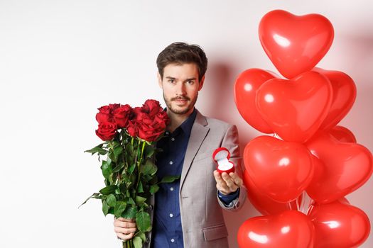 Handsome man in suit giving an engagement ring and bouquet of red roses, marry me on Valentines day, standing with heart balloons on white background
