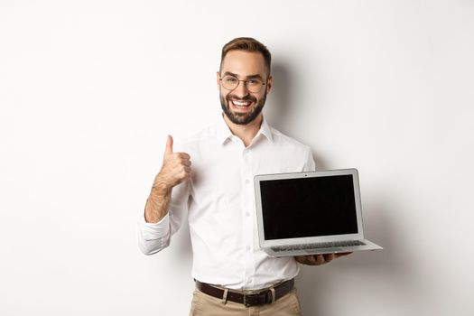 Successful business man showing laptop screen, make thumb up in approval, praise something good, standing over white background