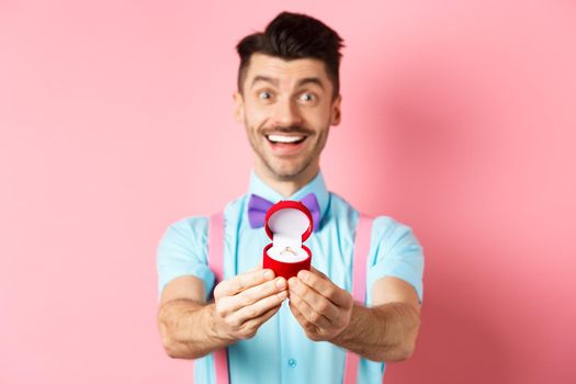 Valentines day. Romantic young man in bow-tie stretch hands with engagement ring and smiling, asking to marry him, making proposal, standing over pink background