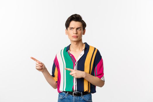 Suspicious young man looking and pointing fingers left with doubtful face, standing unsure in summer shirt, white background