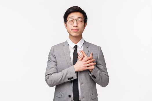 Waist-up portrait of romantic handsome young asian male entrepreneur, office manager have crush on his coworker, hold hands on heart close eyes, daydreaming about falling in love, white background