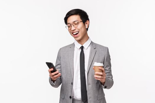 Office workers, business and lifestyle concept. Waist-up portrait of lucky cheerful asian male entrepreneur calling friend using wireless earphones, hold smartphone and take-away coffee, smiling.