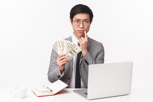 Waist-up portrait of thoughtful, serious-looking smart young asian businessman holding huge sum of money, squinting puzzled and touch lip, pondering how invest money, sit at office with laptop