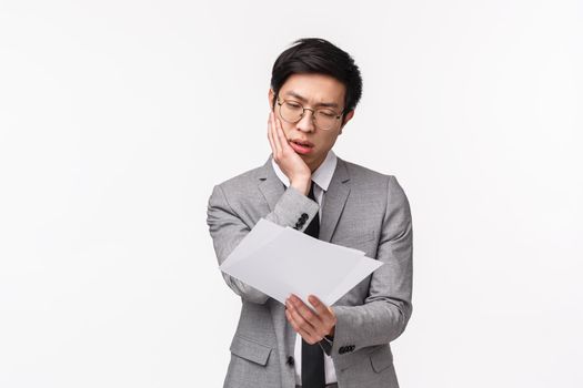 Waist-up portrait of shocked and displeased young asian businessman reading disappointing report, looking at document with sighing moody expression, standing white background sad
