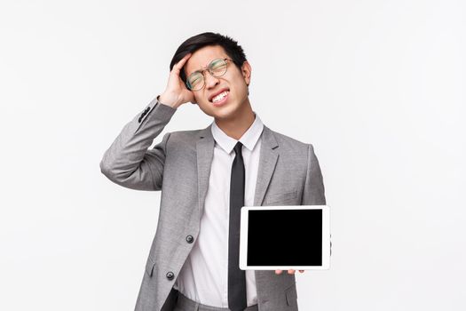 Waist-up portrait of embarrassed or forgetful young asian businessman forgot something, remember about important thing, showing digital tablet screen, grimacing bothered and scratch head