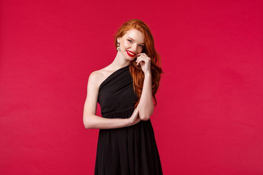 Elegance, fashion and woman concept. Portrait of seducative and sensual woman with ginger hair in elegant evening black dress, attend prom or party, wearing red lipstick, smiling coquettish