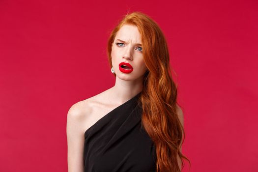 Close-up portrait of perplexed and confused young redhead woman seeing something strange, squinting and frowning as looking left troubled identify what is this, stand red background