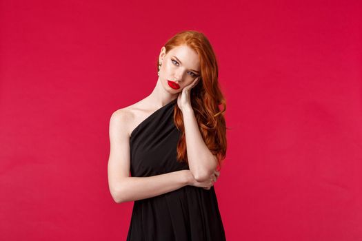 Portrait of annoyed and bored reluctant young redhead female in black evening dress, tired of people, boring party or prom night, lean on palm look camera gloomy, stand red background