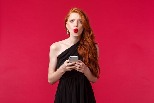 Portrait of concerned and confused young redhead woman being dumped by message on her prom night, look frustrated dont understand what happened, wear black dress, holding smartphone