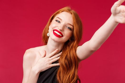 Close-up portrait of gorgeous smiling, happy redhead woman enjoying her birthday party, taking selfie for social media in stylish black dress and prom look with red lipstick, red background