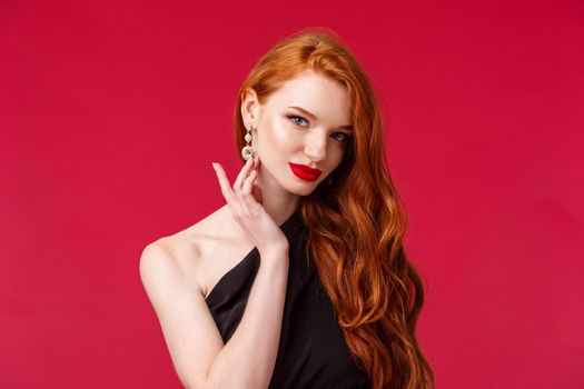 Close-up portrait of sensual seductive redhead woman with red lipstick, touching earring coquettish look camera, slightly smiling as flirting with person on party, wear black dress, red background