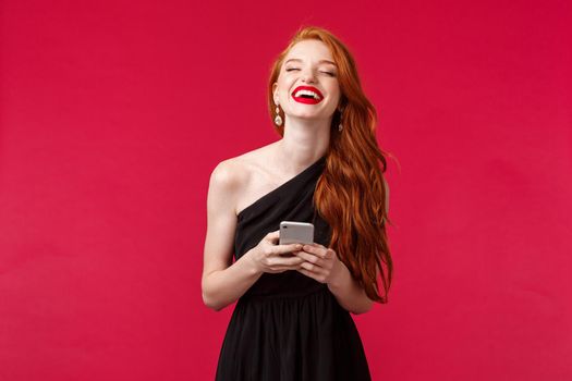 Technology and lifestyle concept. Gorgeous young redhead woman in black dress laughing over red background, holding smartphone, texting friend during prom discuss funny moment
