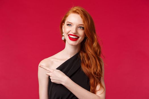 Close-up portrait of cheerful enjoyable young redhead girl in red lipstick, black dress, pointing and looking left with pleased intrigued expression, see something interesting, advertisement concept
