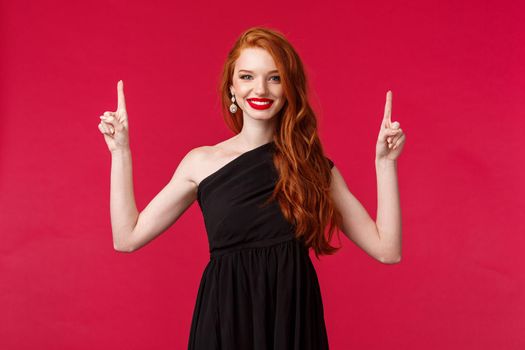 Celebration, events, fashion concept. Portrait of confident charming redhead female in elegant black dress, evening makeup, showing good store for buying prom outfit, pointing fingers up, smile