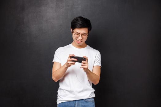 Technology, gadgets and people concept. Portrait of concentrated, focused asian guy trying kill mob in mobile game, passing arcade or street races, holding smartphone horizontally and grimacing