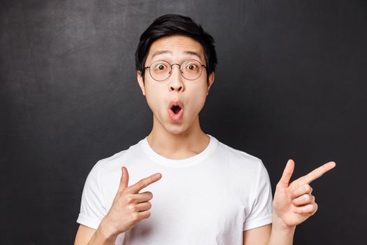 Close-up portrait of thrilled and amused asian young man in white t-shirt, open mouth amazed say wow, pointing fingers right at something impressive and stunning, white background