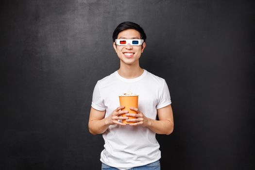 Leisure, movies and lifestyle concept. Handsome cheerful and excited asian man in 3d glasses, holding popcorn and smiling amused as watching favorite thriller or comedy, black background