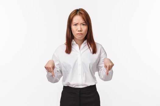 Portrait of angry grumpy asian female entrepreneur scolding employee, pointing down and frowning camera, pouting upset, look here gesture as being disappointed with bad project, white background.