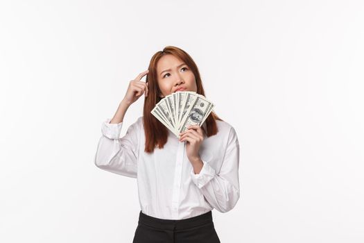 Portrait of indecisive and thoughtful young successful asian woman earn money and dont know how invest them, scratching head uncertain smirk pondering, holding fan of cash, white background