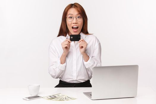 Career, work and women entrepreneurs concept. Close-up portrait cheerful young asian woman sitting in office with laptop, money, hold credit card, look amused, got first paycheck