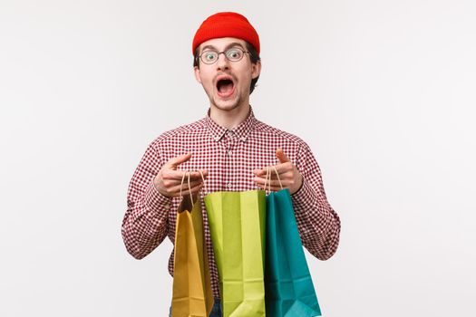 Waist-up portrait of funny and cute caucasian guy in glasses, red beanie, buy lots staff for girlfriend, hope she will like it, holding shopping bags and staring camera, wasted all paycheck in stores
