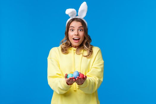 Amused cute and lovely caucasian blond woman in rabbit ears holding Easter eggs, give it to family member as celebrating divine holiday together, standing blue background