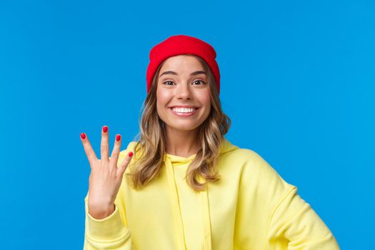 Happy smiling female student in hipster red beanie, showing number four, fourth and grinning at camera optimistic, make reservation or order, standing blue background upbeat
