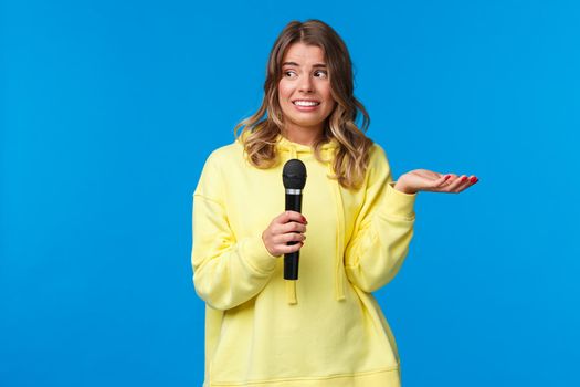 Awkward cute blond girl dont know lyrics as singing karaoke, looking at friend nervous smile, embarrassed sing in fron of people, hold microphone, standing blue background