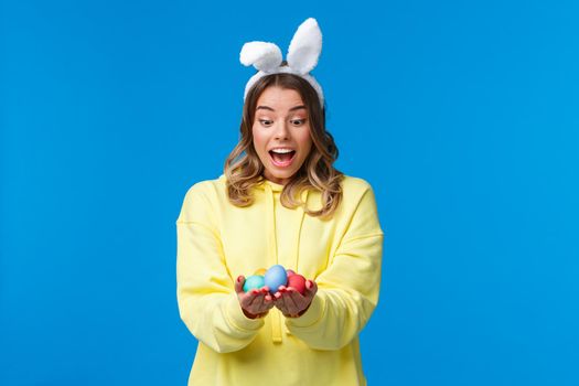 Surprised emotive cute blond caucasian girl playing holiday game, found Easter eggs look amused, wearing rabbit ears as celebrating religious holiday on sunday, blue background