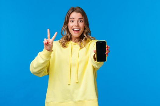 Optimistic cute blond european girl with short haircut, yellow hoodie, showing kawaii peace gesture and mobile phone display as using photo filter to edit and post pic online, blue background