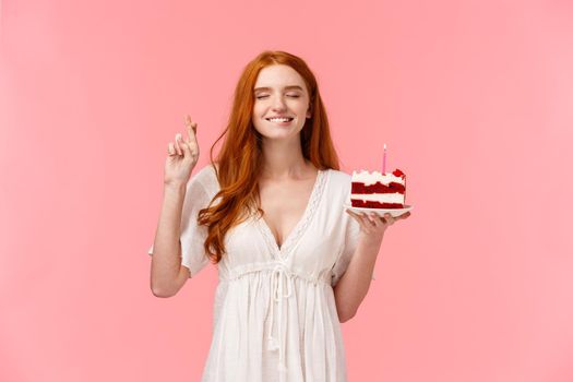 Celebration, happiness concept. Attractive dreamy redhead girl in lovely white dress, holding b-day cake, cross finger good luck, bite lip tempting close eyes and making wish to blow candle