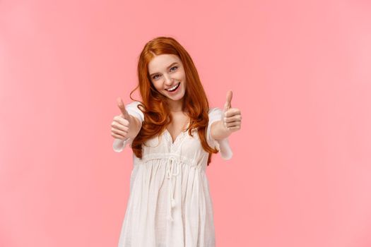 I support your choice. Charismatic excited, happy redhead woman in white dress, showing thumbs-up to cheer you, agree or express satisfaction of cool product, smile approval, pink background