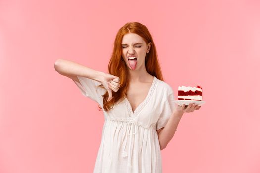 Ignorant, picky and snobbish redhead birthday girl hate b-day cake, holding plate with dessert, showing tongue grimacing and make thumb-down in disapproval, aversion and disgust