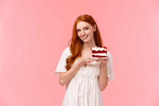 Lovely, romantic and alluring redhead girlfriend baked delicious surprise for valentines day date, holding peace cake on plate and smiling tempting with sassy, coquettish expression, pink background