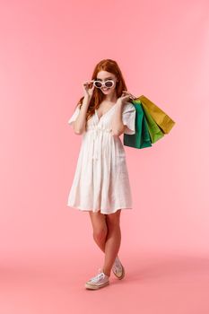Full-length vertical shot alluring happy redhead female release stress with shopping, shopaholic walking along stores, shop-malls with bags behind back, take-off glasses, smiling camera