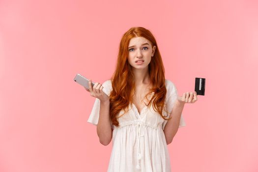 Indecisive and unsure cute european redhead woman shrugging, looking confused and useless camera, shrugging with smartphone and credit card in hands, dont know what order online