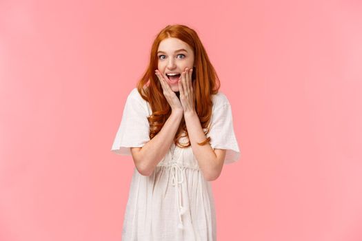 Expectation, anticipation and happiness concept. Amused cute redhead silly girl looking surprised and excited, gasping fascinated, looking charmed and amazed camera, pink background