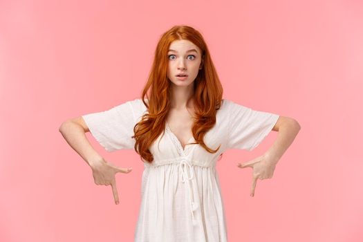 Curious and intrigued redhead girl heard interesting news, raise eyebrows in wonder and amazement, pointing fingers down as asking question about product, stand pink background