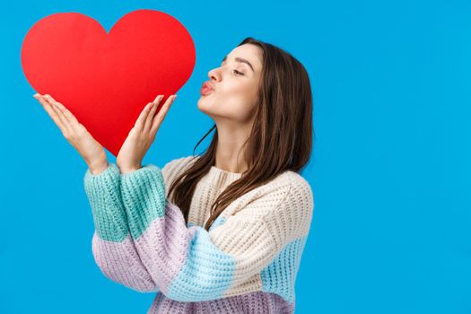 Dreamy young woman cherish her relationship, prepare valentines day gift, kissing big cute red heart sign over left side copy space, standing blue background delighted and upbeat