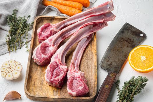 Raw lamb chops or mutton cuts, with ingredients carrot orange, herbs, and old butcher cleaver knife, on white stone background