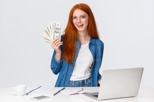 Girl likes her new work, got promoted and receive first paycheck. Attractive sassy redhead woman in glasses, standing near working table, laptop, holding money, big cash