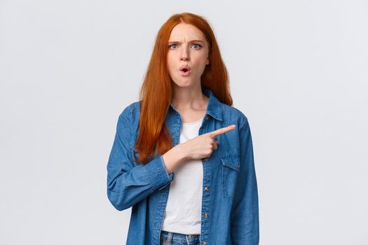 What is that. Displeased, tensed and mad frustrated redhead bossy woman pointing right waiting explanation as staring puzzled and upset, standing white background bothered